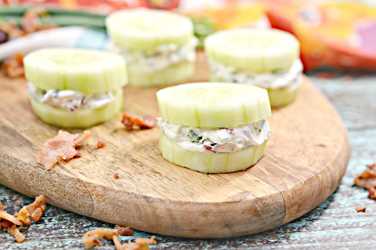 Four cucumber sandwich bites filled with a creamy mixture are arranged on a wooden serving board. The filling appears to have herbs and possibly bits of bacon. There are a few scattered pieces of bacon on the board, with a colorful background slightly blurred.