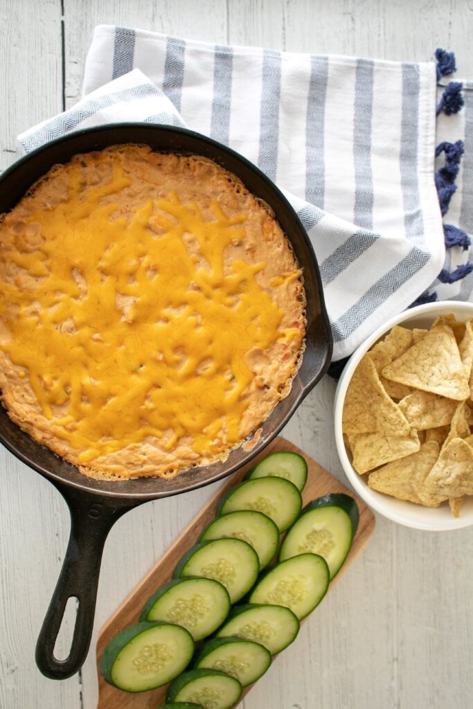 A cast iron skillet filled with a baked cheesy dip is placed on a cloth napkin. Next to it, a bowl of tortilla chips and a wooden cutting board with sliced cucumbers are arranged on a white wooden table.