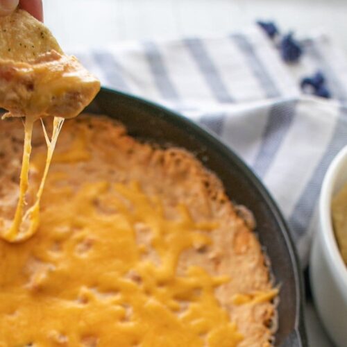 A close-up of a hand holding a tortilla chip with melted cheese dip from a pan. The cheese stretches from the dip to the chip. The pan of hot cheese dip is on a blue and white striped cloth, with a bowl of tortilla chips next to it.