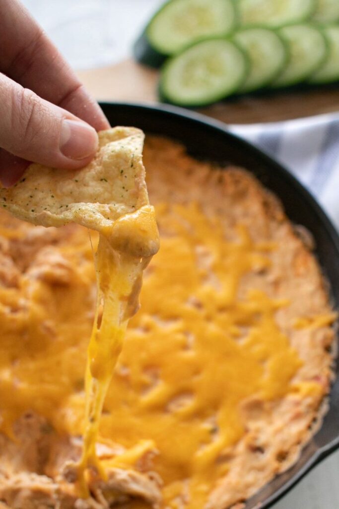 A close-up of a person dipping a tortilla chip into a cheesy dip in a cast iron skillet. The cheese is melted and stretchy. In the background, there are sliced cucumbers on a wooden cutting board.