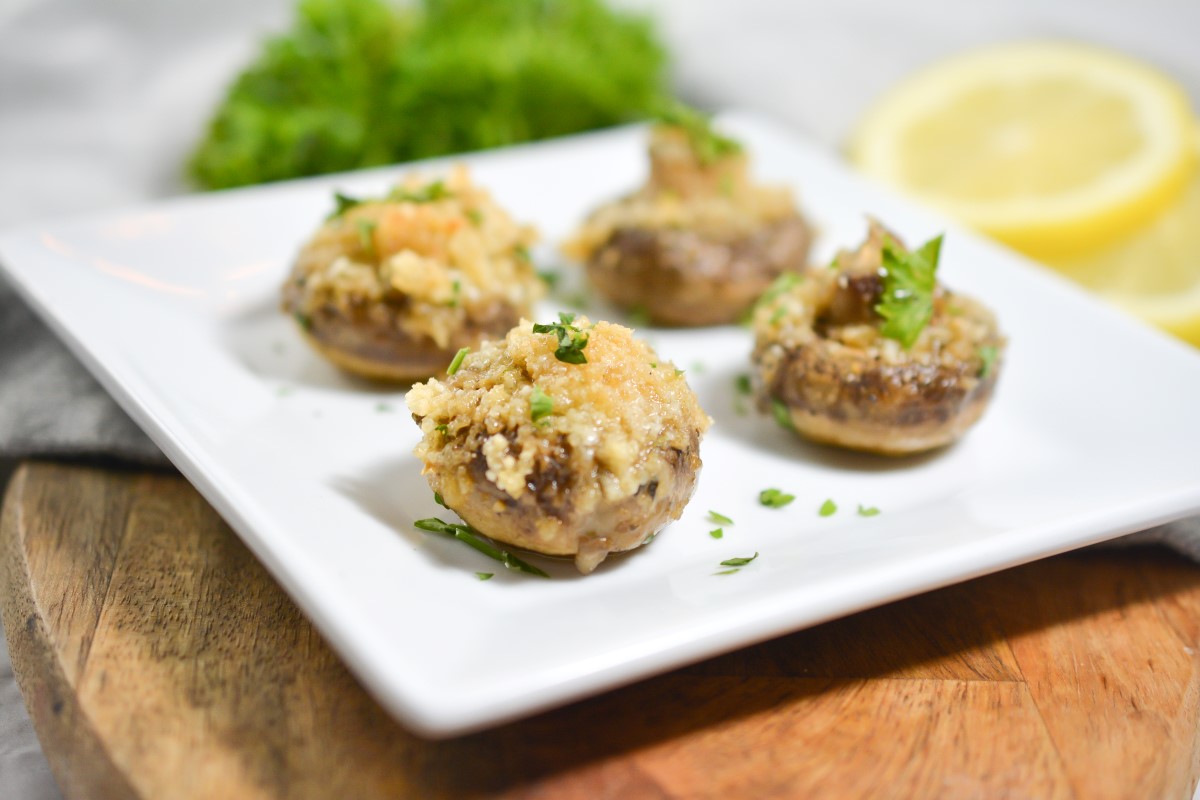 A white square plate holds four stuffed mushrooms garnished with chopped parsley. The mushrooms are placed on a wooden board. In the background, there is a leafy green garnish and lemon slices blurred out of focus.
