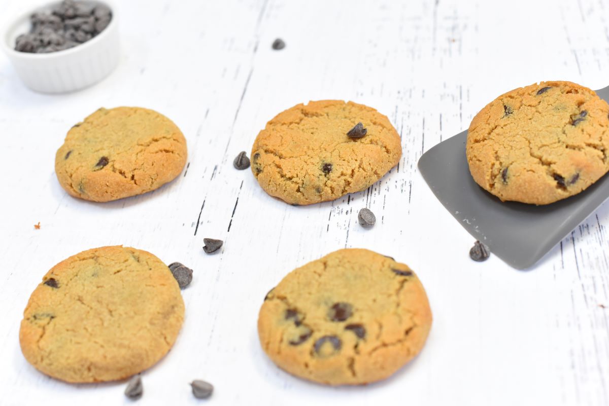 Five chocolate chip cookies on a white wooden surface. Four cookies are directly on the surface, and one cookie is being lifted by a grey spatula. A small white bowl containing chocolate chips is at the top left corner of the image.