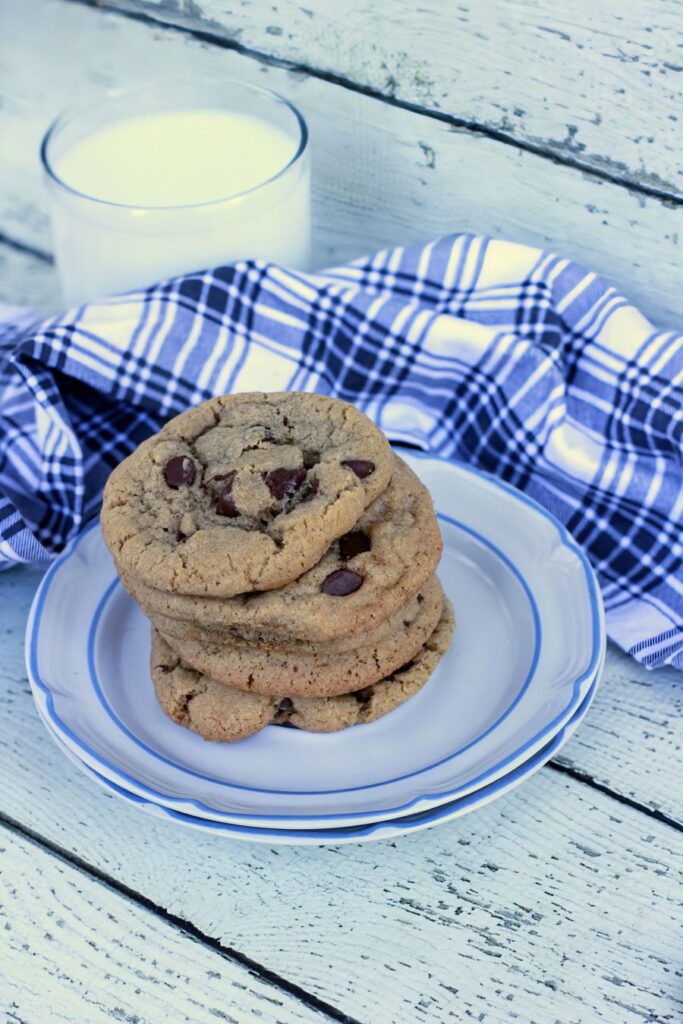 A stack of gluten-free chocolate chip cookies on a white plate with blue trim. A blue and white plaid cloth is draped beside the plate, and a glass of milk sits in the background on a rustic white wooden surface.