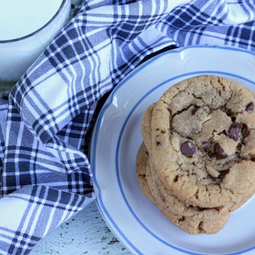 A stack of gluten-free chocolate chip cookies sits on a white plate with a blue rim. Next to the plate is a glass of milk and a blue and white checkered cloth, all arranged on a white wooden surface.