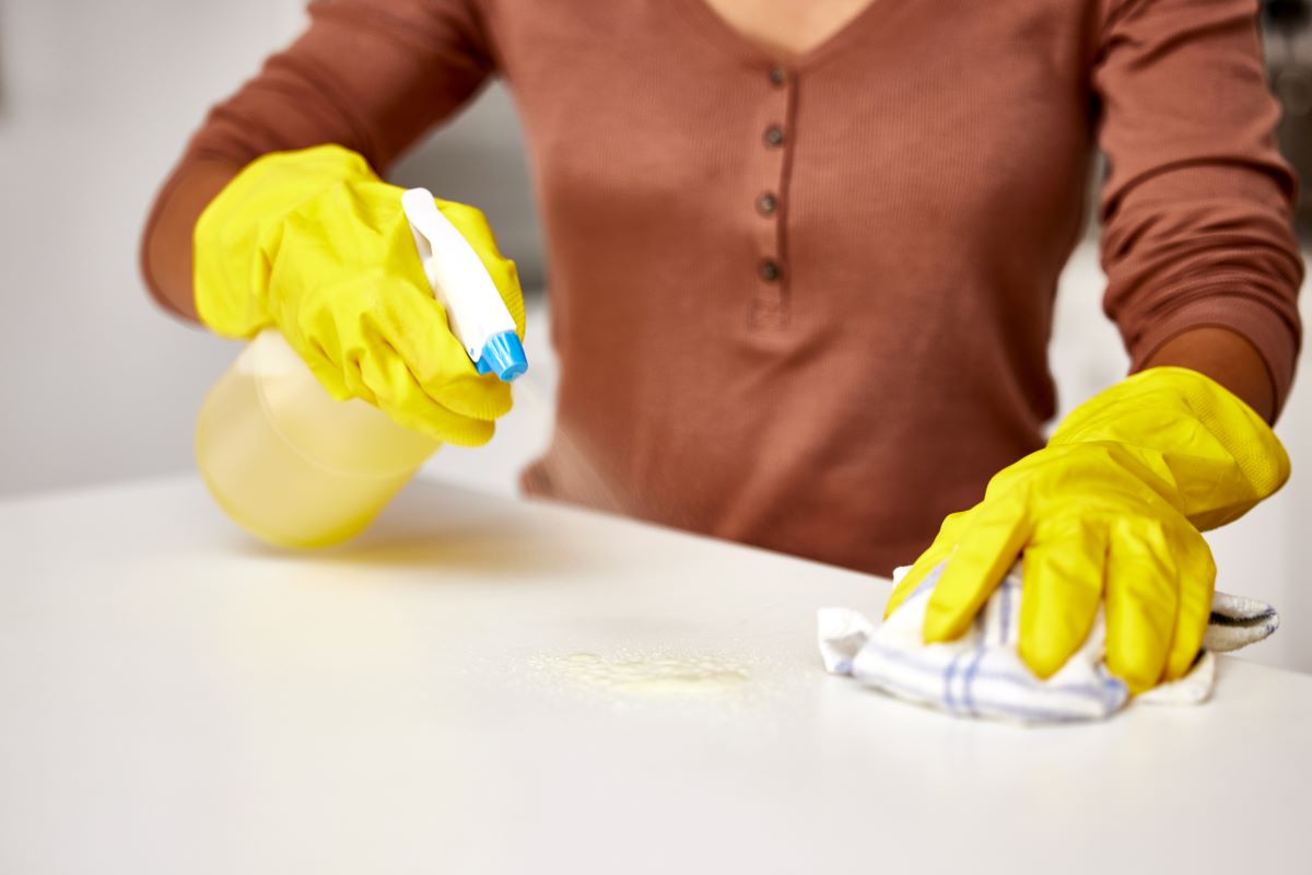 Person wearing yellow gloves cleaning a surface with a spray bottle in one hand and a cloth in the other. The focus is on the cleaning process. The person is wearing a brown top.