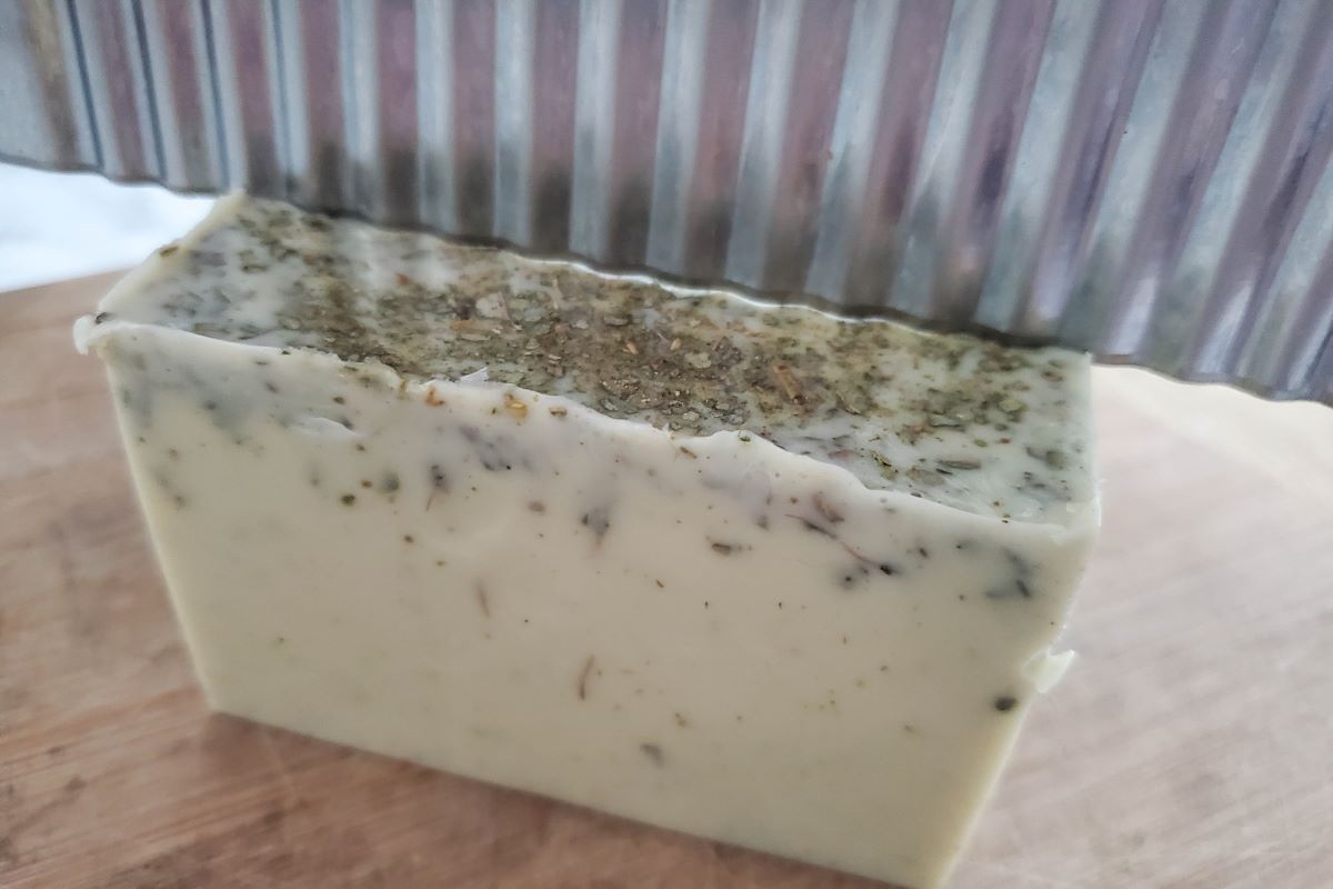 A bar of homemade soap with specks, being sliced with a wavy cutter on a wooden surface.
