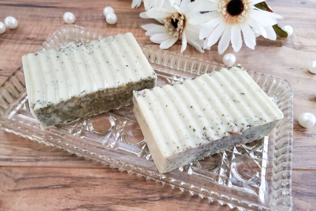 Two bars of handmade soap with a ribbed pattern are displayed on a clear, decorative glass tray. The soap contains dark specks and is placed on a wooden surface. White daisies and small decorative pearls surround the tray, adding a natural touch.