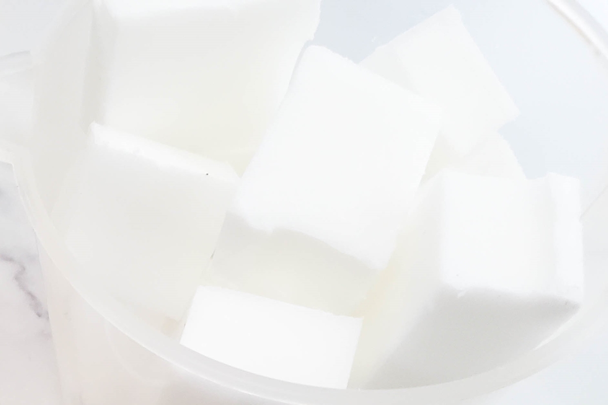 A close-up image of a white container filled with white, soap-like cubes. The cubes have a clean, solid appearance and are evenly cut, stacked together in the container. The background is a soft white, enhancing the minimalistic look.