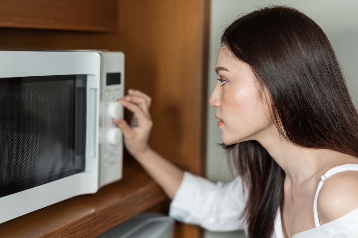 A woman with long dark hair adjusts the settings on a microwave oven. She is wearing a white top and is focused on the task. The microwave is positioned in a wooden cabinet.