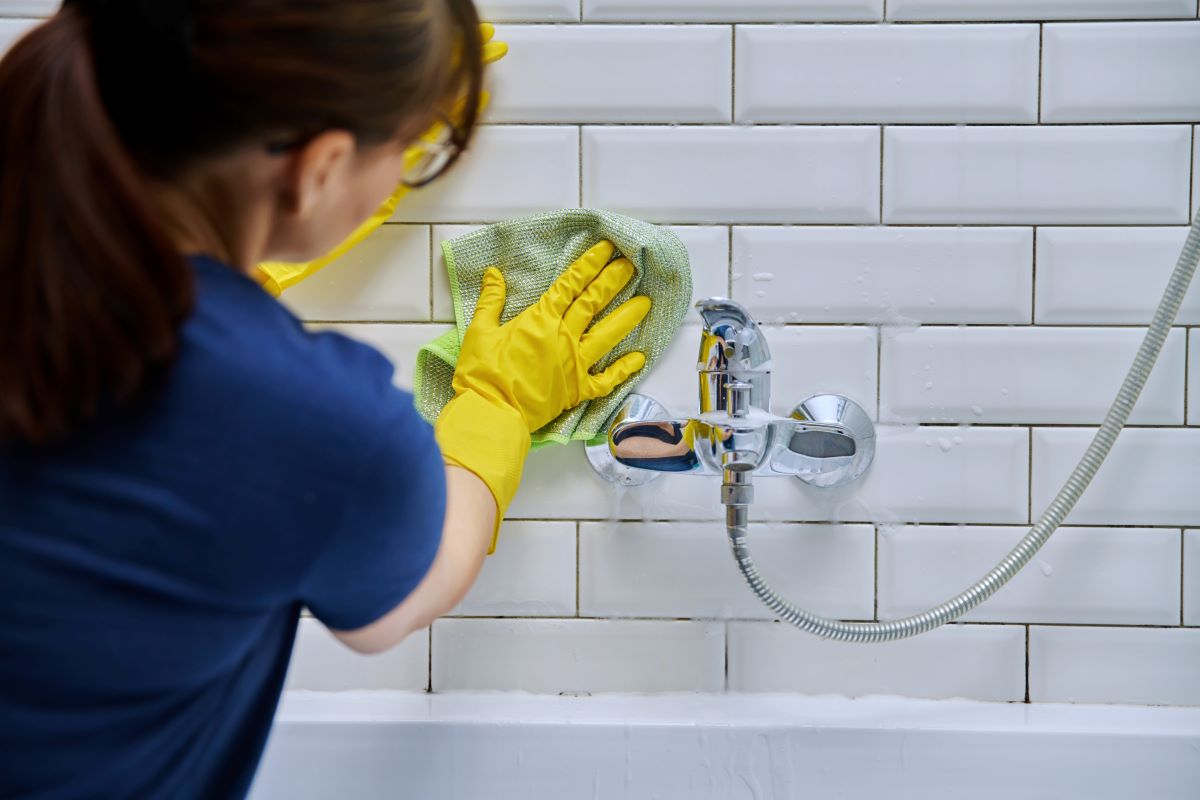 A person wearing a navy shirt and yellow rubber gloves cleans white tiled bathroom walls with a green cloth. A silver faucet and shower hose are visible. The setting is bright and tidy.