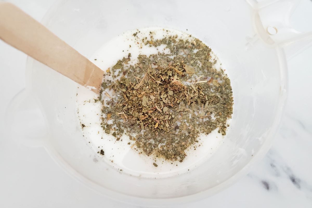 A clear plastic cup filled with white liquid and topped with dried herbs, with a wooden stick submerged in the mixture, resting on a white marble surface.