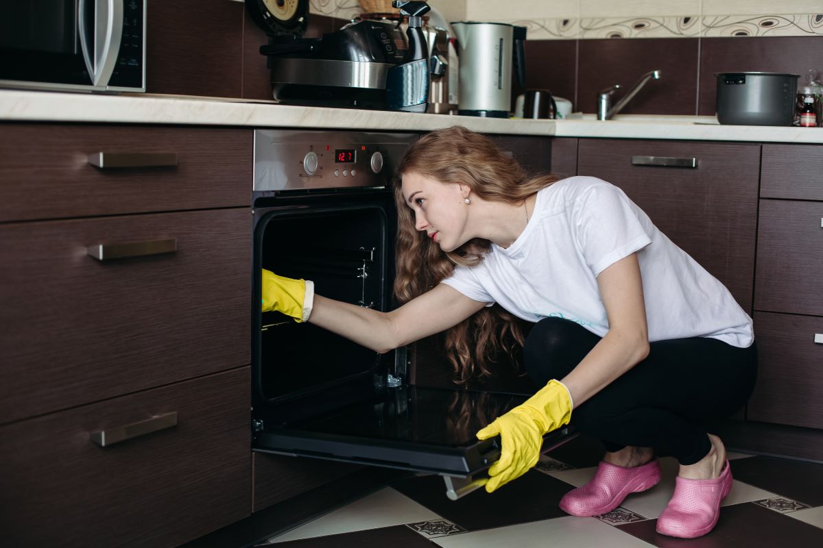 A woman wearing yellow gloves and pink shoes is crouching and cleaning the inside of an open oven in a modern kitchen.