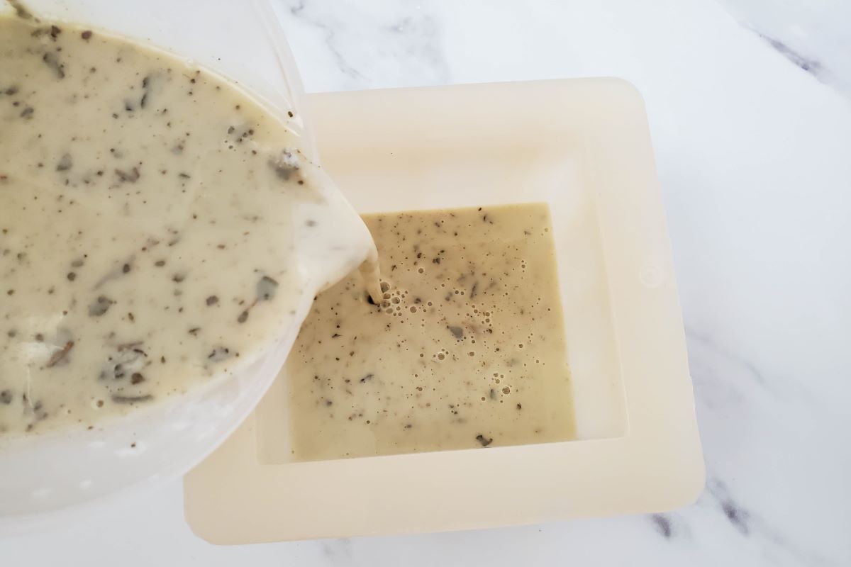 A creamy liquid with herbs is being poured from a transparent pitcher into a square pale mold, set on a white marble surface.