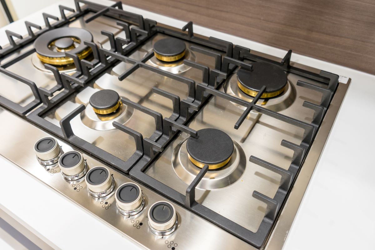 A stainless steel gas stovetop with five burners and black grates, featuring control knobs at the front. The stovetop is set against a wooden backsplash.
