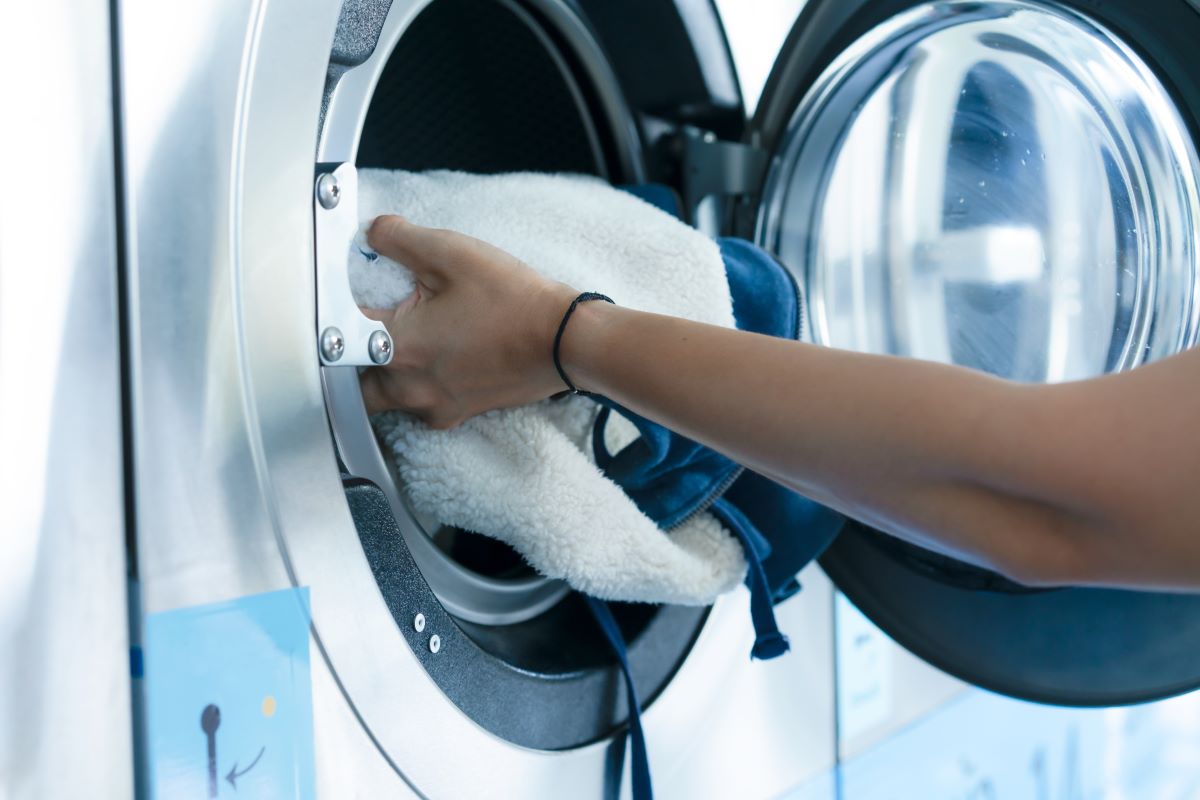 A person's arm places a fluffy white blanket into a front-loading washing machine. The machine is stainless steel, and the door is open. A paper with a design is partially visible below the opening.