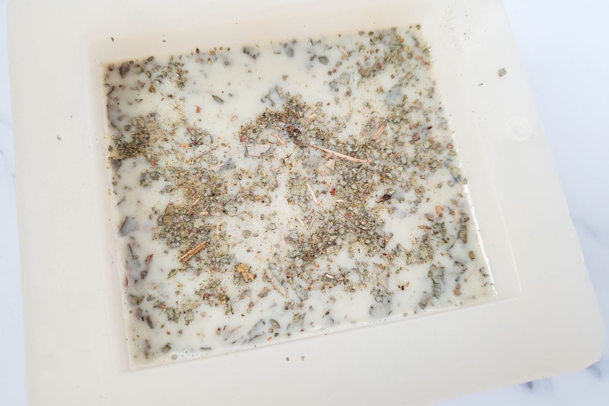 A close-up of a square soap mold containing a mixture of white soap base with various herbs and small particles scattered on top, giving a textured appearance. The mold rests on a light surface.
