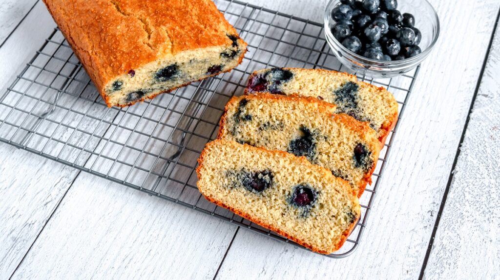 A loaf of blueberry bread rests on a cooling rack atop a wooden table. Three slices of the bread are cut and displayed. A small glass bowl filled with fresh blueberries is visible in the background.