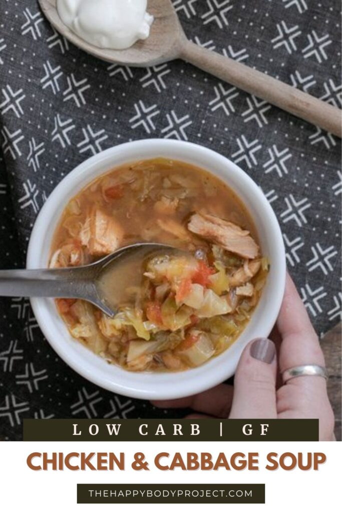A bowl of chicken and cabbage soup on a patterned cloth. A hand with silver rings holds a spoon in the soup. Above is a wooden spoon with sour cream. Text at the bottom reads "Low Carb | GF Chicken & Cabbage Soup TheHappyBodyProject.com".