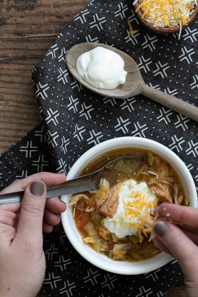 A person adding shredded cheese to a bowl of soup topped with a dollop of sour cream. A wooden spoon with more sour cream rests nearby on a patterned black and white cloth. The soup appears to contain meat and vegetables.