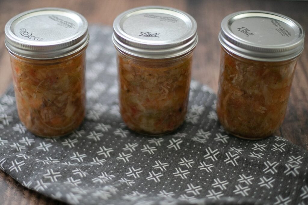 Three glass jars with metal lids are filled with homemade sauerkraut. They are placed on a patterned cloth on a wooden surface.