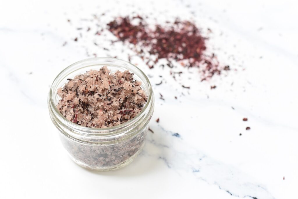 A small glass jar filled with coarse, pinkish-purple sea salt, placed on a white surface. Additional salt and seasoning are scattered in the background, adding texture and color contrast.
