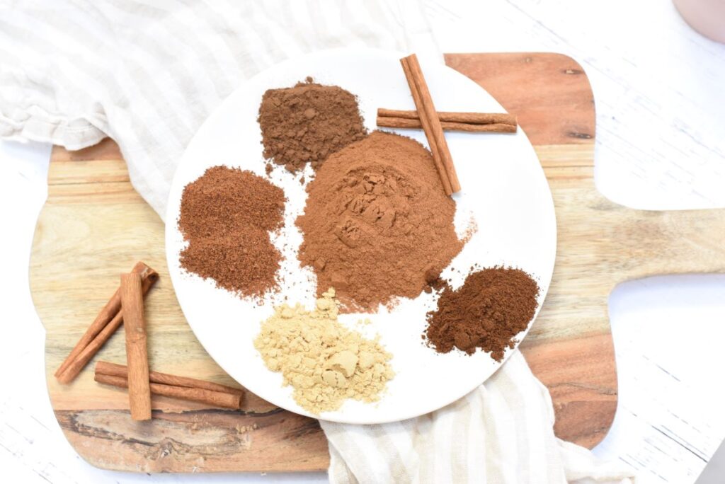 A white plate on a wooden board holds various brown and beige spice powders, including cinnamon sticks, ginger, and other spices. A striped cloth is partially visible beneath the plate.