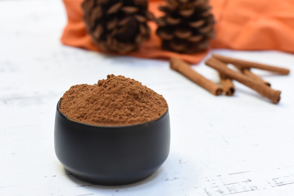 A black bowl filled with homemade spices is placed on a white surface. In the background, there are two pine cones, an orange cloth, and a few cinnamon sticks.