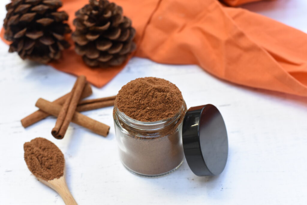 A black bowl filled with homemade spices is placed on a white surface. In the background, there are two pine cones, an orange cloth, and a few cinnamon sticks.