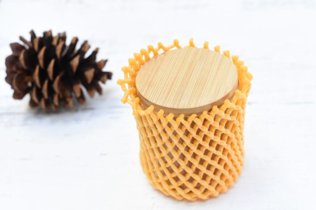 A wooden container wrapped in a protective orange mesh sits on a white surface. A pinecone is placed nearby, adding a natural element to the composition.