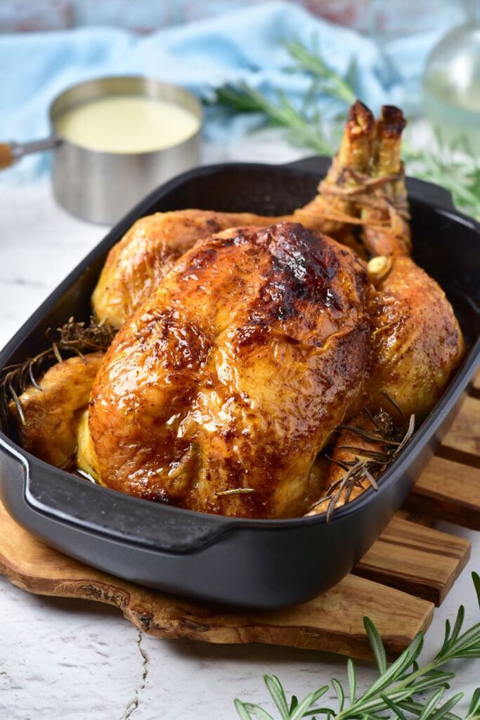 A golden-brown roasted chicken sits in a black roasting pan, garnished with rosemary sprigs. A creamy sauce in a small pan is visible in the background, along with a blue cloth. The scene rests on a wooden cutting board.