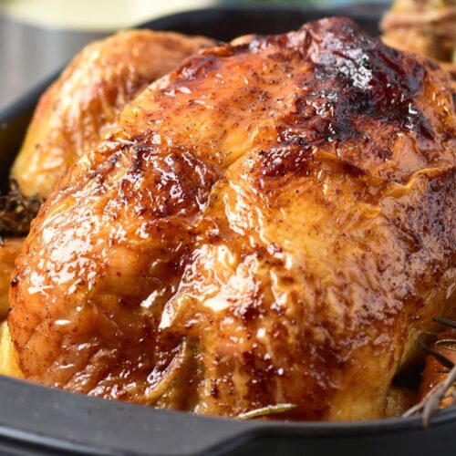 A close-up of a perfectly roasted chicken with golden, crispy skin in a black roasting pan. Visible are sprigs of rosemary, enhancing the presentation. The chicken looks juicy and appetizing, ready to be served.