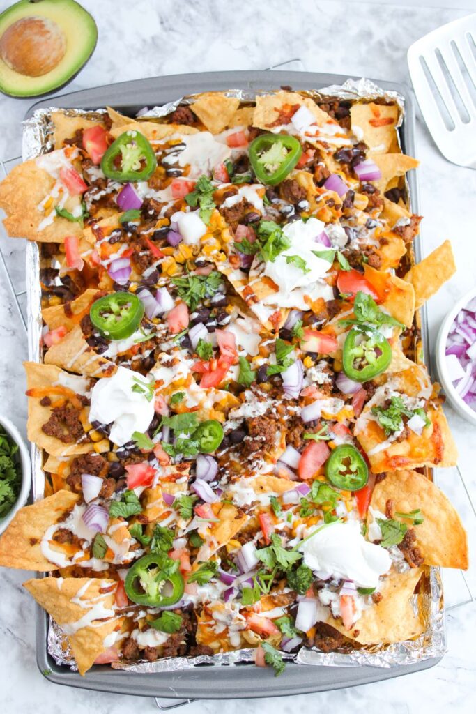 A tray of loaded nachos topped with melted cheese, ground meat, diced tomatoes, red onions, jalapeño slices, cilantro, and dollops of sour cream. An avocado half and a spatula are visible nearby on a marble surface.