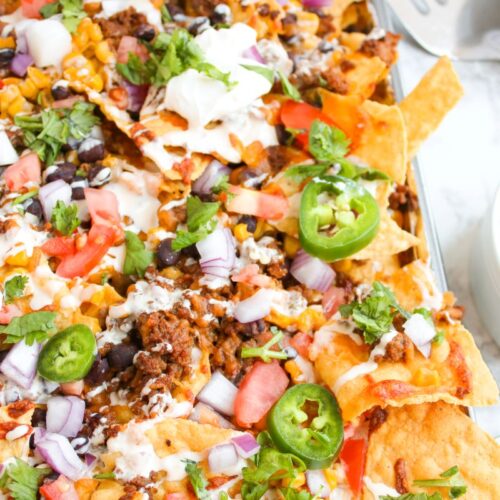 A close-up of loaded nachos topped with ground meat, cheese, jalapeño slices, sour cream, chopped onions, diced tomatoes, black beans, corn, and cilantro. A bowl of chopped red onions is on the side.