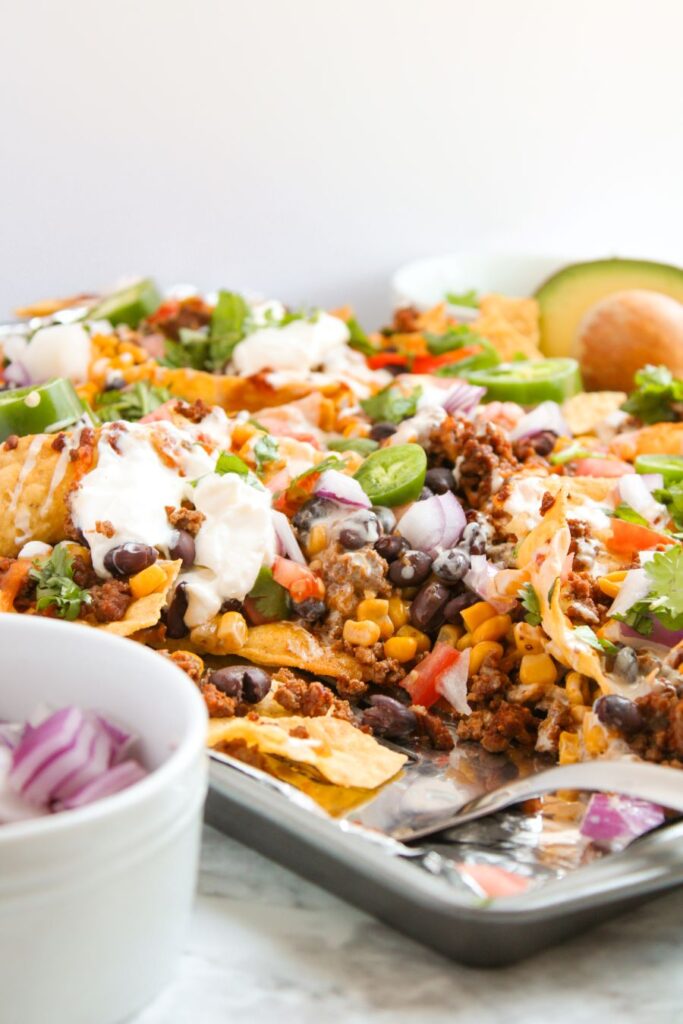 A tray of loaded nachos topped with ground meat, melted cheese, black beans, corn, jalapeños, chopped onions, tomatoes, cilantro, and dollops of sour cream. A bowl of chopped red onions and a sliced avocado are nearby.