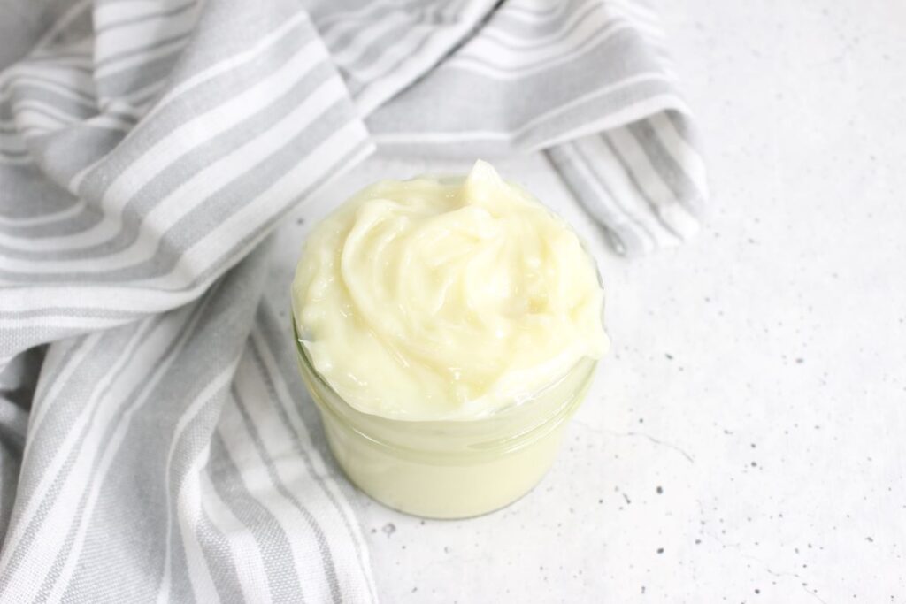 A jar filled with creamy, white mayonnaise is placed on a light-colored countertop. A gray and white striped cloth is casually draped beside the jar.