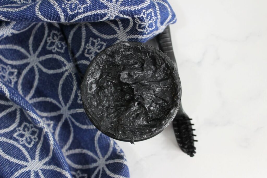 An open jar of activated charcoal toothpaste sits beside a black toothbrush on a blue and white patterned cloth. The background features a light-colored surface, enhancing the sleek, modern feel of this dental duo.