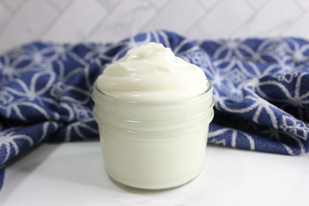 A small glass jar filled with creamy white lotion sits on a white surface. Behind it, a blue cloth with a floral pattern is slightly blurred, creating a soft background.