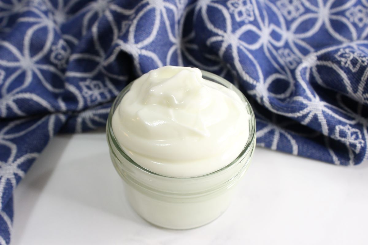 A small glass jar filled with creamy white yogurt sits on a white surface. In the background, a blue and white patterned cloth is partially visible.