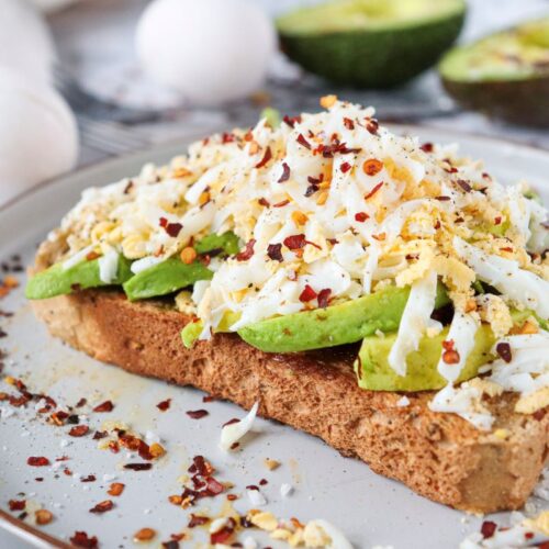 A slice of toast topped with sliced avocado and shredded egg, sprinkled with red pepper flakes and spices. In the background, there are two halves of an avocado and two whole eggs on a marble countertop.