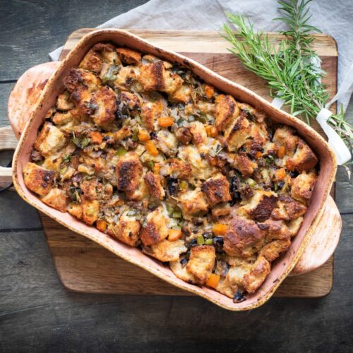 A baked stuffing dish with golden bread cubes, herbs, and vegetables in a wooden baking dish on a cutting board. Fresh rosemary lies beside it, and a white ribbon is placed on a wooden table underneath.