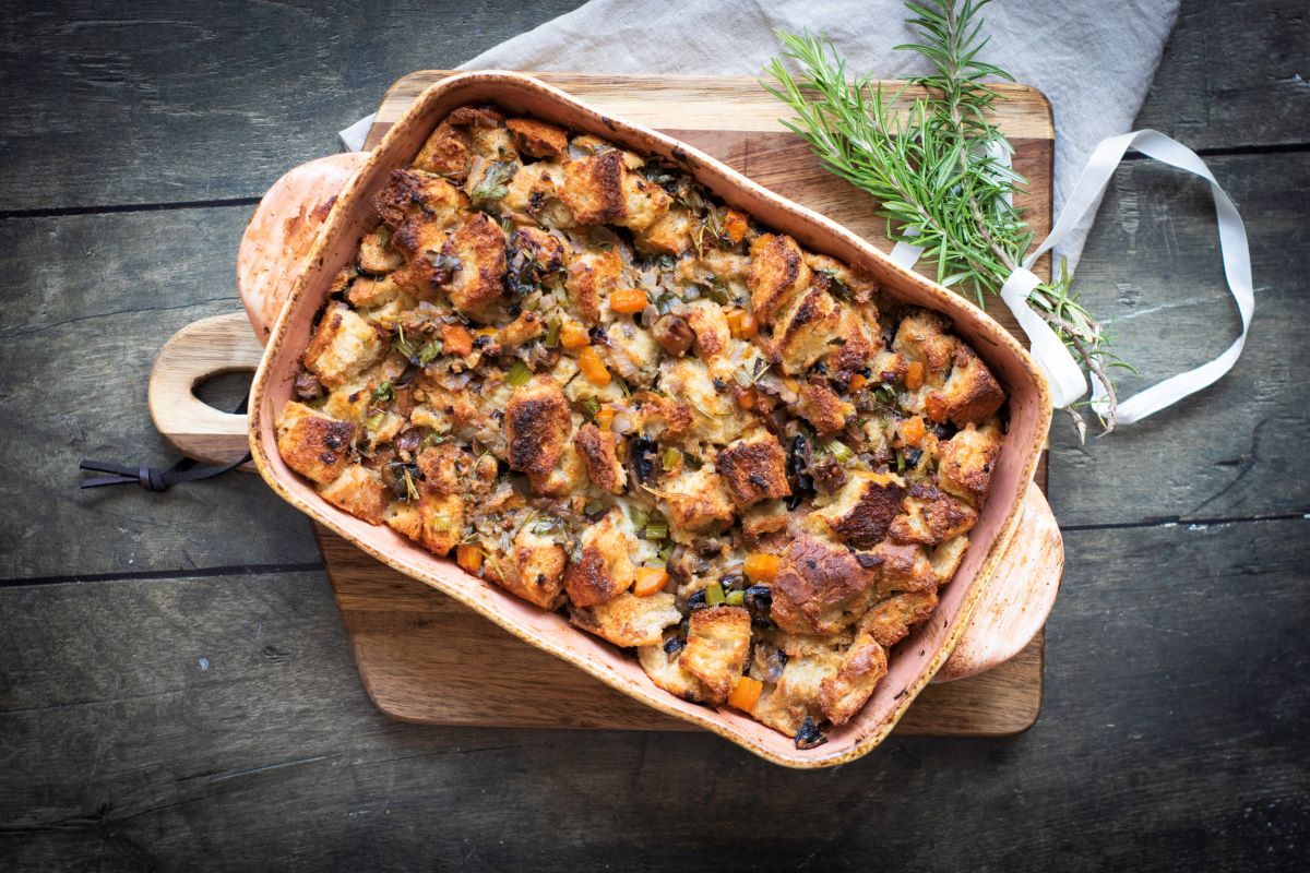 A baked stuffing dish with golden bread cubes, herbs, and vegetables in a wooden baking dish on a cutting board. Fresh rosemary lies beside it, and a white ribbon is placed on a wooden table underneath.