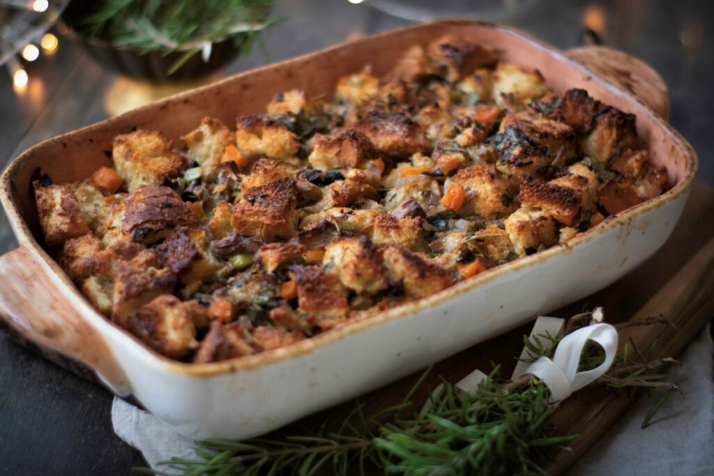 A baked casserole dish filled with golden-brown stuffing, featuring chunks of bread, chopped vegetables, and herbs. It's placed on a wooden board, garnished with sprigs of rosemary nearby.