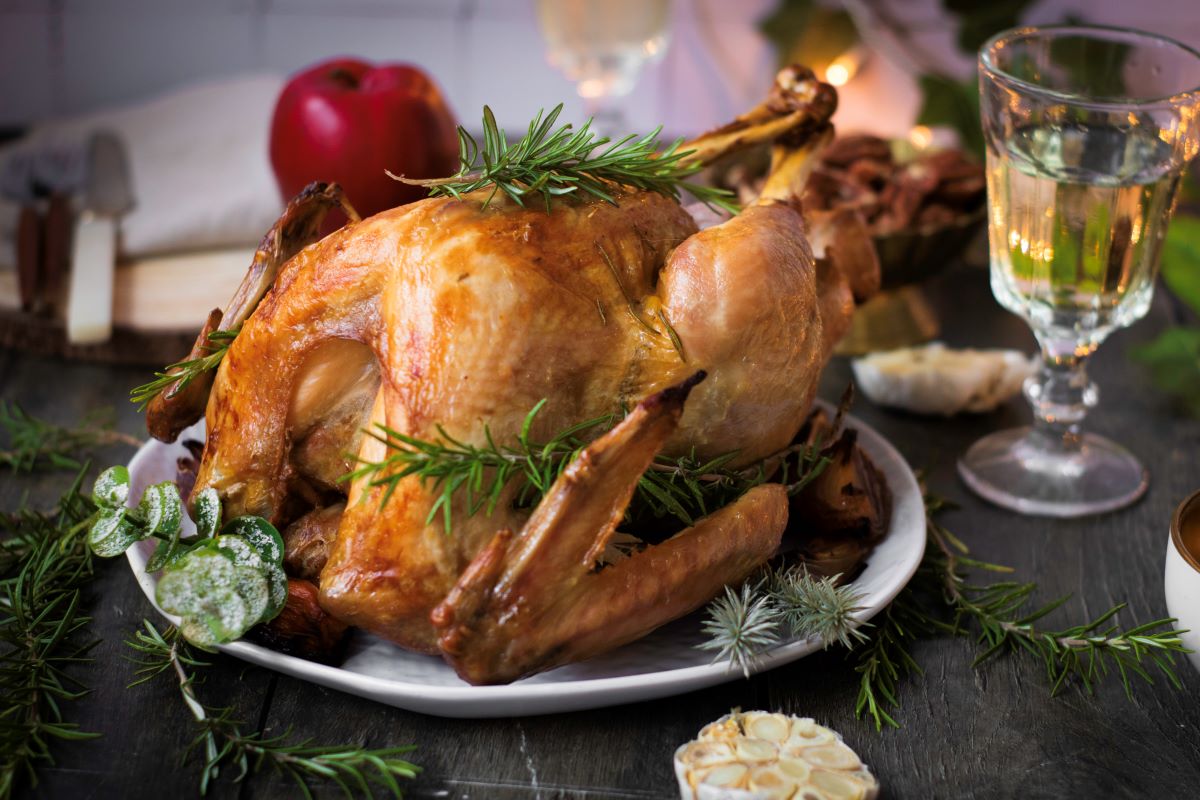 A golden-brown roasted turkey garnished with fresh rosemary on a white platter. In the background, there's a glass of white wine, a red apple, and a few greens scattered around. .