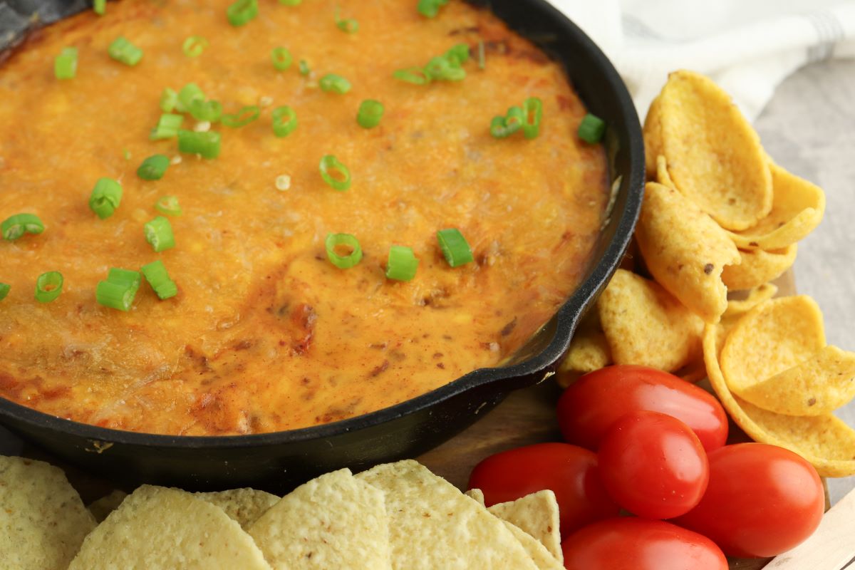A skillet filled with melted cheesy dip topped with chopped green onions. Surrounding the skillet are tortilla chips, scoop chips, and a bunch of cherry tomatoes.