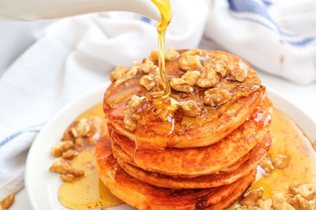 A stack of fluffy pancakes topped with walnuts is being drizzled with syrup. The pancakes are arranged on a white plate, with a white and blue napkin in the background. The syrup creates a glossy, appetizing look.