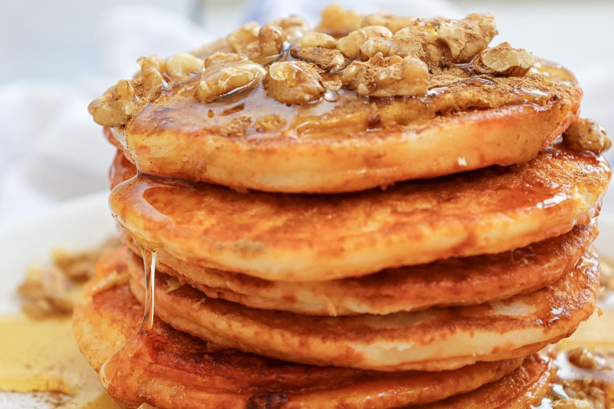 A close-up of a stack of golden-brown pancakes topped with walnuts and drizzled with syrup, served on a white plate.