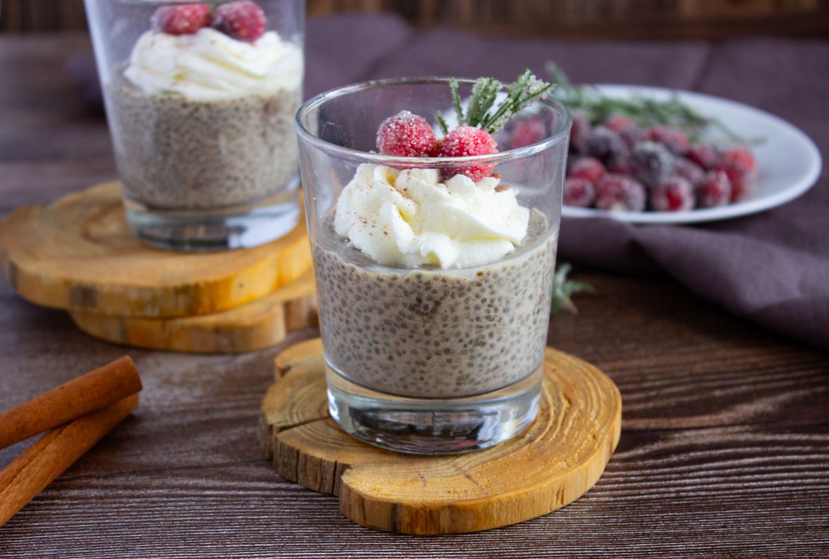 Two glasses of chia pudding topped with whipped cream and sugared cranberries. Each glass rests on a wooden coaster. In the background, a plate with additional cranberries and a brown cloth are visible. Cinnamon sticks are on the table.