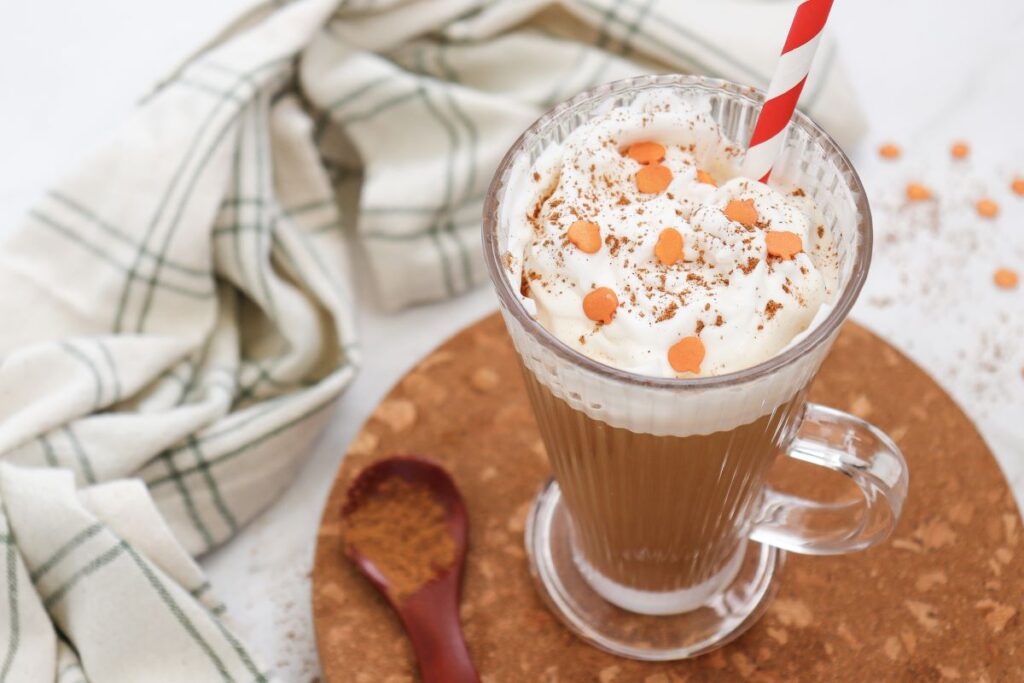 A glass mug filled with a creamy coffee drink topped with whipped cream, sprinkled with orange chips and cinnamon. A red and white striped straw is inserted. A striped cloth napkin and a wooden spoon with brown powder are nearby.