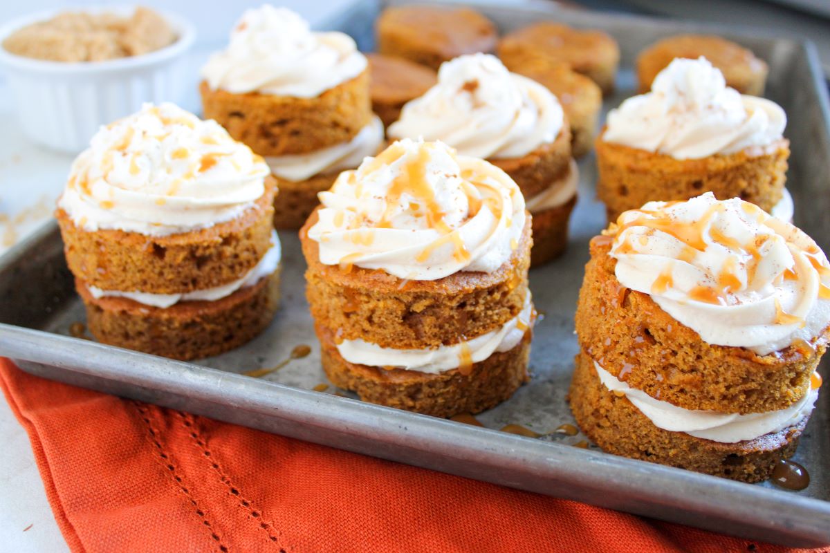 Mini pumpkin cakes with whipped cream and caramel drizzle are stacked on a metal tray. The cakes are layered with cream and rest on an orange cloth. A bowl of brown sugar is visible in the background.