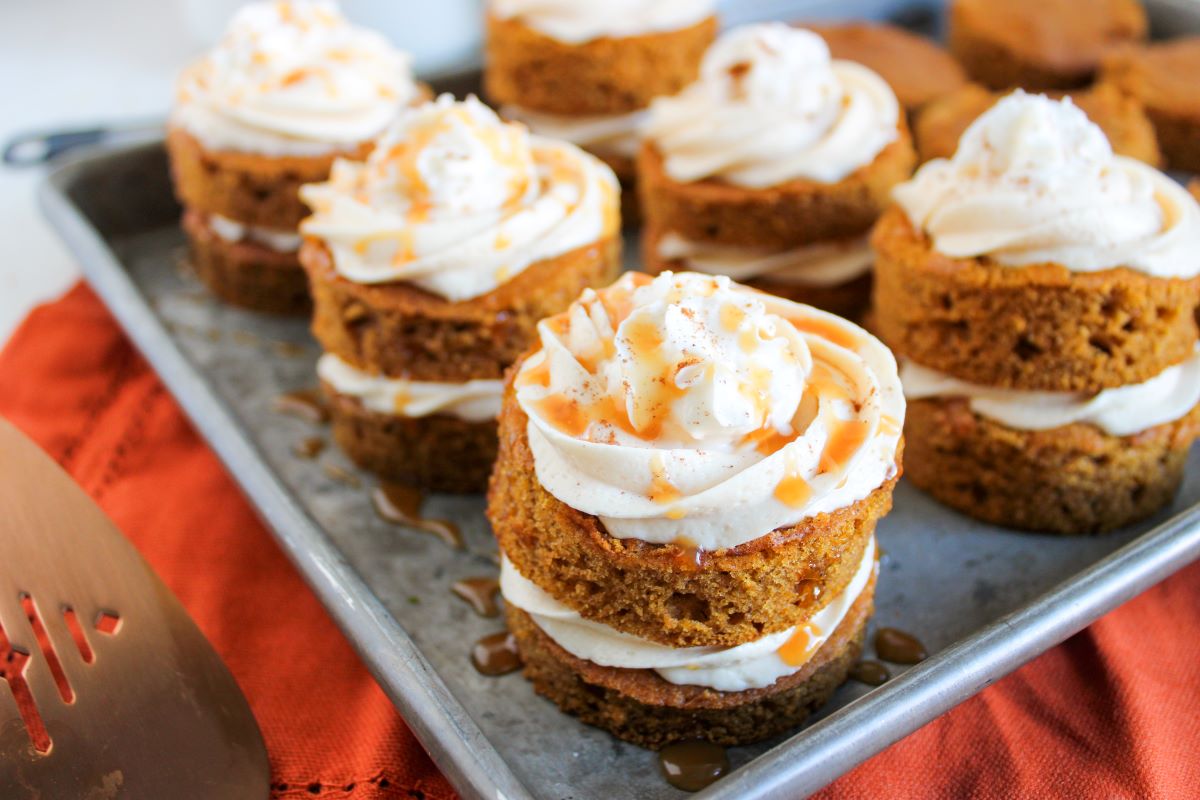 Mini pumpkin cakes stacked in layers with creamy white frosting, drizzled with caramel sauce, arranged on a metal tray with an orange cloth underneath.
